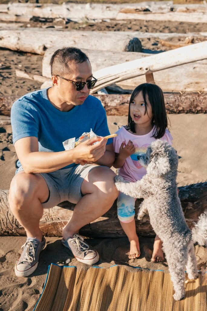 A father, daughter, and their dog having a fun snack time on a sunny beach.